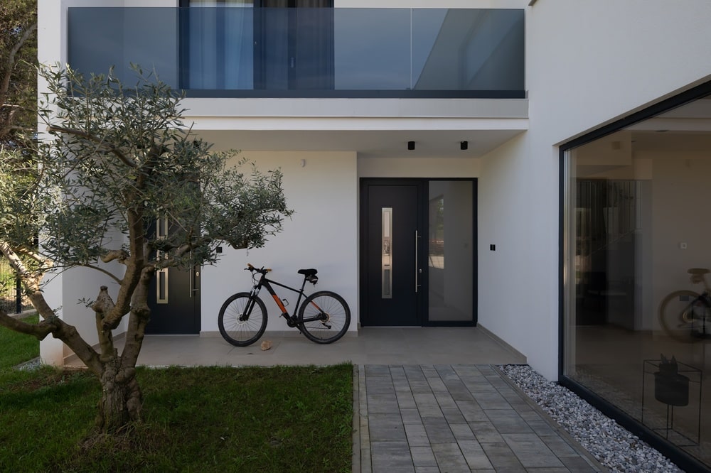 Architecture Moderne : Entrée de Maison avec Vélo et Olivier Entrée d'une maison moderne blanche avec vélo noir, porte sombre, olivier et balcon en verre fumé.