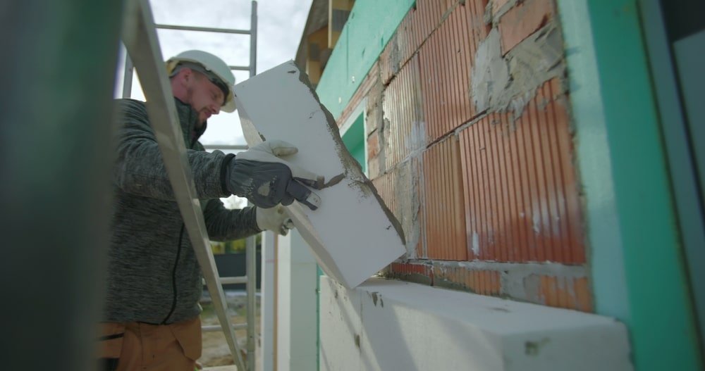 Ouvrier en casque blanc installant un panneau d'isolation thermique par l'extérieur sur un mur en briques.