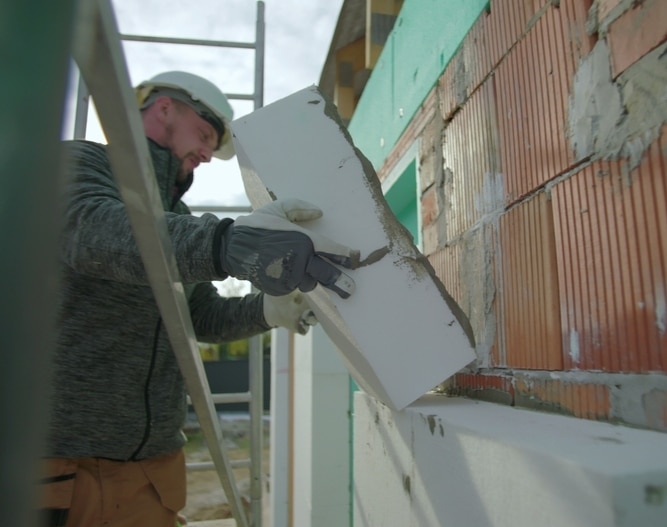 Isolation Thermique par l'Extérieur d'un Bâtiment Ouvrier installant un panneau d'isolation blanc sur un mur en briques pour améliorer l'efficacité énergétique.