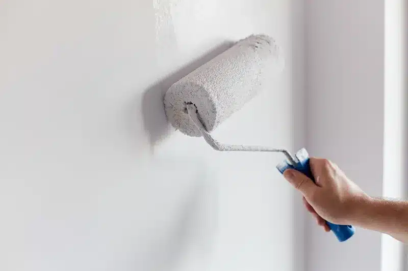 Peinture blanche au rouleau : réussir vos rénovations Main tenant un rouleau de peinture blanc appliquant une couche fraîche sur un mur pour une rénovation intérieure.